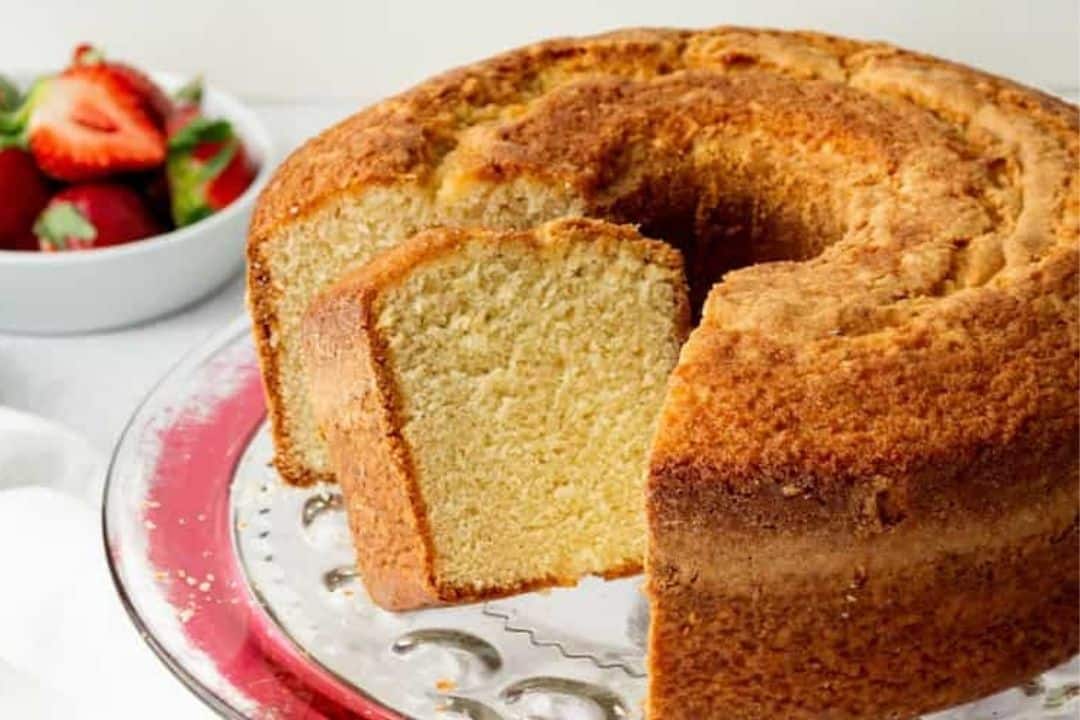 A sliced bundt pound cake on a glass cake stand with strawberries in a bowl in the background.