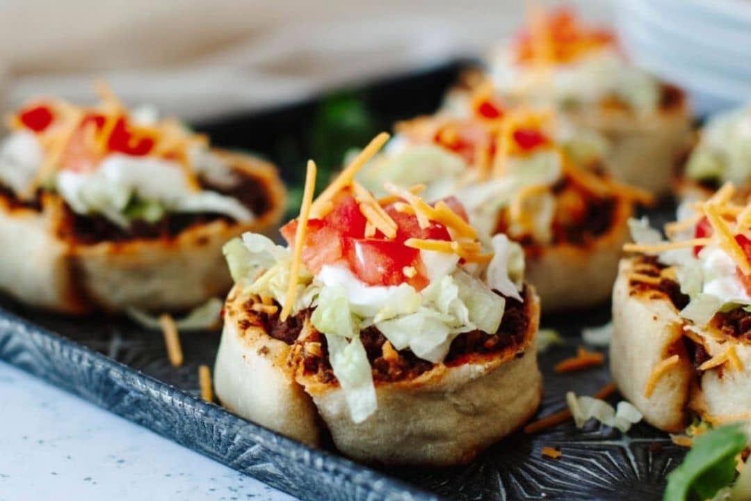 Mini taco cups filled with ground beef, lettuce, tomatoes, cheese, and sour cream on a gray tray.