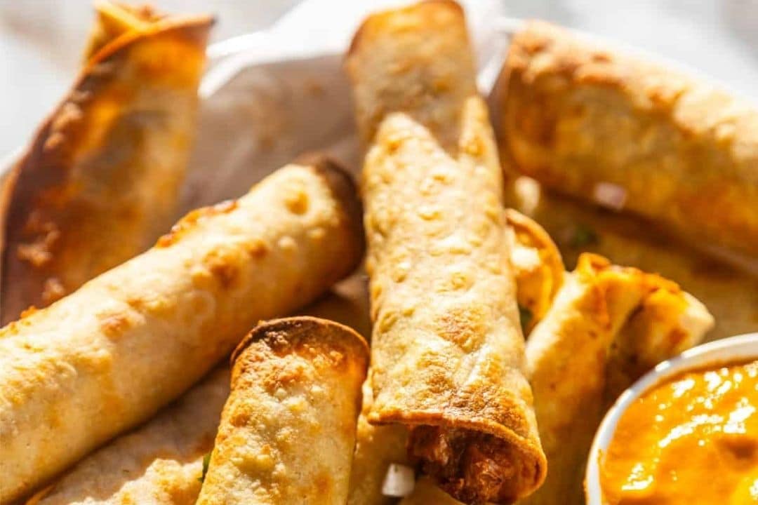 Crispy golden taquitos stacked next to a small bowl of orange dipping sauce.