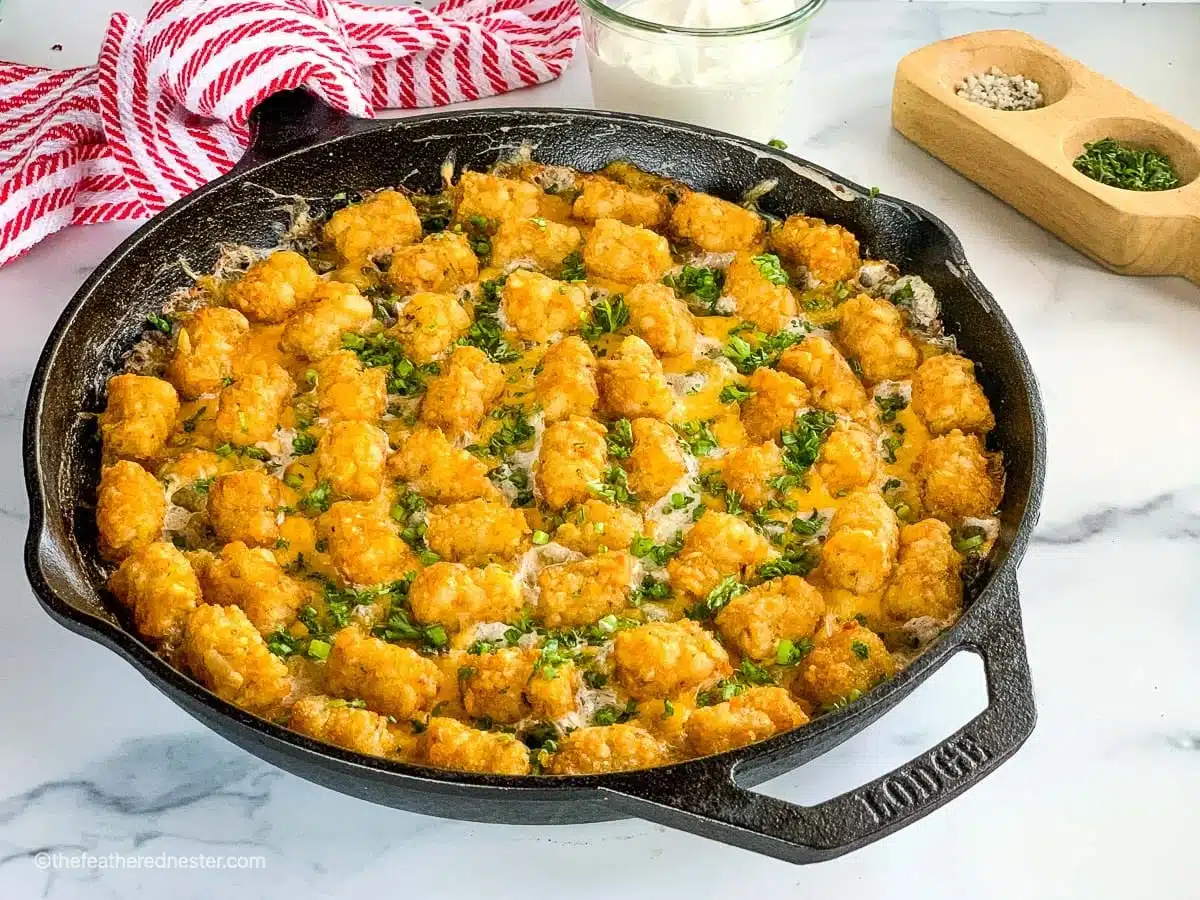 A skillet filled with tater tot casserole, topped with golden tots and herbs, sits on a marble countertop.