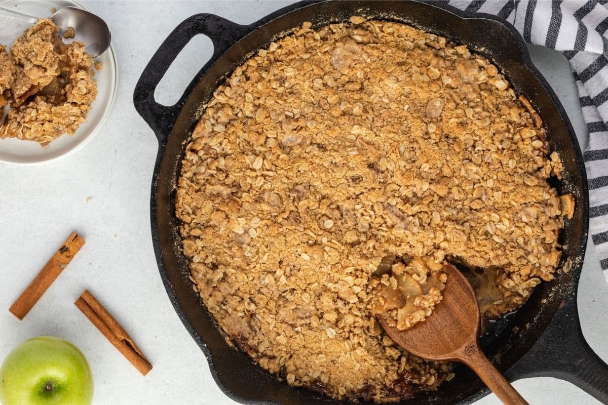 Apple crisp in a cast-iron skillet with a serving on a side plate, two cinnamon sticks, and a green apple.