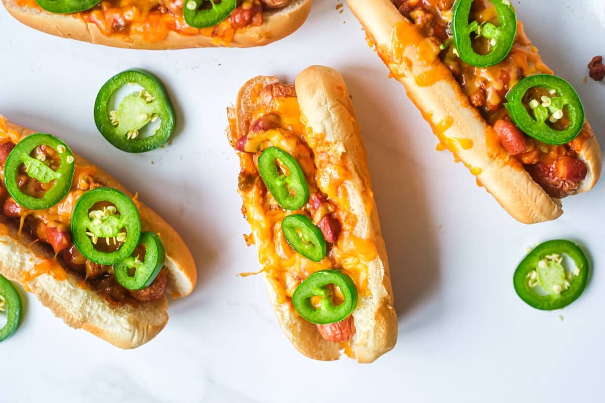 Hot dogs topped with chili, melted cheese, and sliced jalapeños on a white surface.