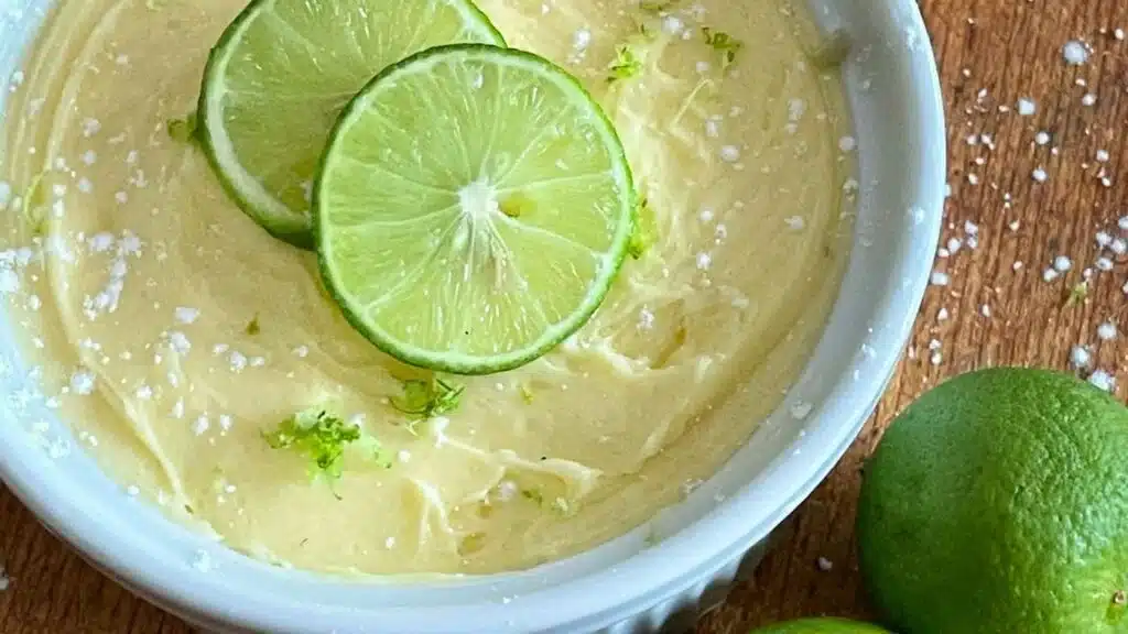 Creamy dessert topped with two lime slices, lime zest, and coarse salt in a white ramekin on a wooden surface.