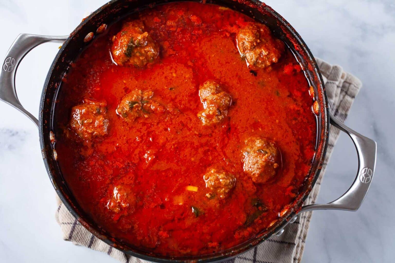 A pot of red tomato sauce with meatballs on a checked kitchen towel, viewed from above.