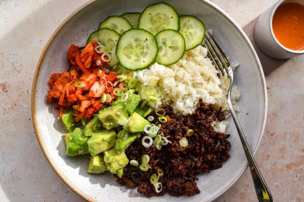 Bowl with rice, ground beef, avocado, kimchi, cucumber slices, green onions, and a fork.