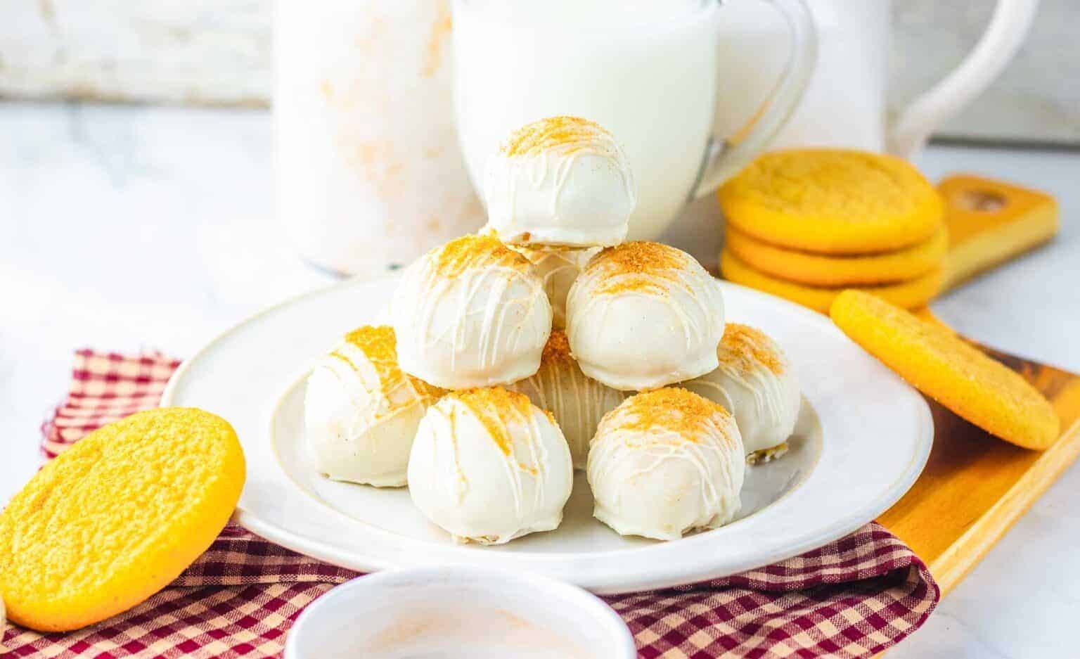 A plate of white chocolate-coated cookies with yellow cookies and a glass of milk in the background.