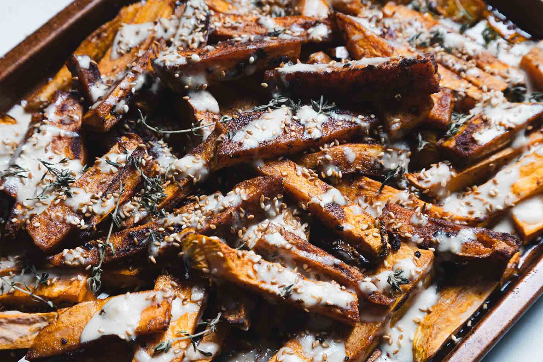 Baked sweet potato fries drizzled with a creamy sauce, sprinkled with seeds and fresh herbs on a tray.