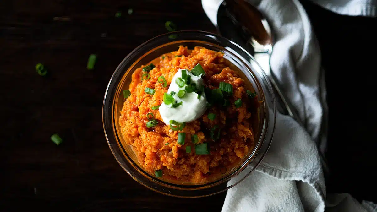 Bowl of mashed sweet potatoes topped with sour cream and chopped green onions, with a spoon and napkin nearby.