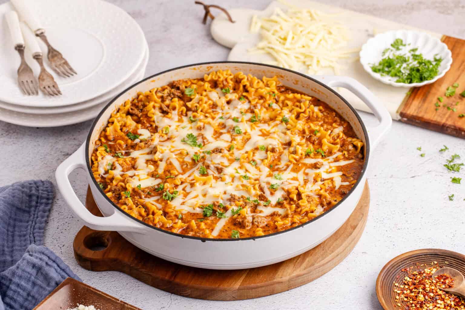 Lasagna soup topped with melted cheese and herbs in a white pot, with plates and shredded cheese nearby.