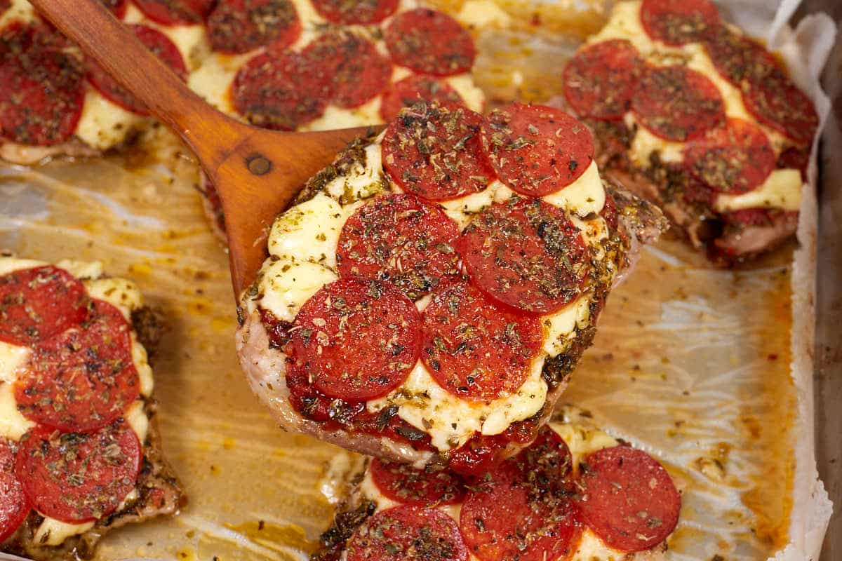 A wooden spoon lifts a piece of pepperoni pizza chicken from a baking tray lined with parchment paper.