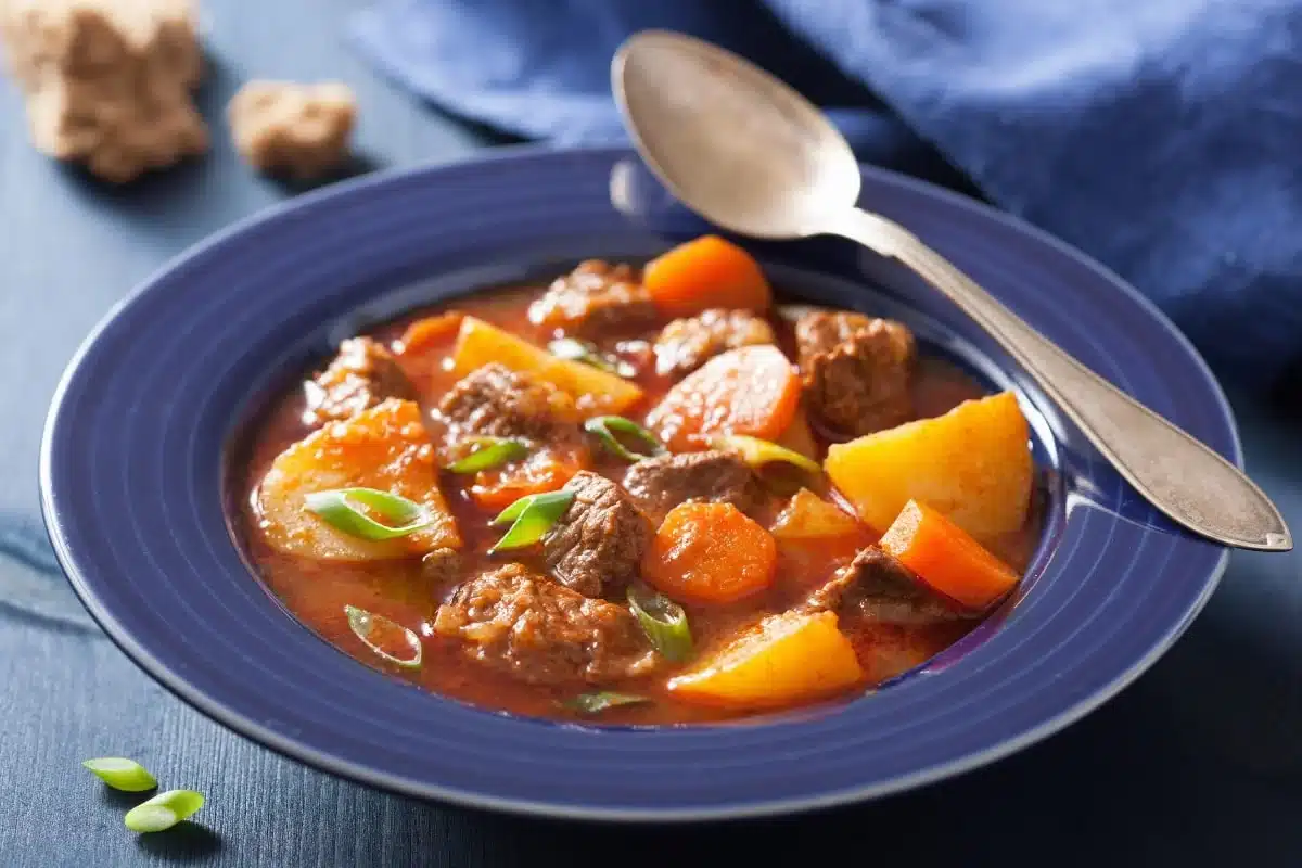 Blue bowl of beef stew with potatoes, carrots, and green onions; a spoon rests on the rim.