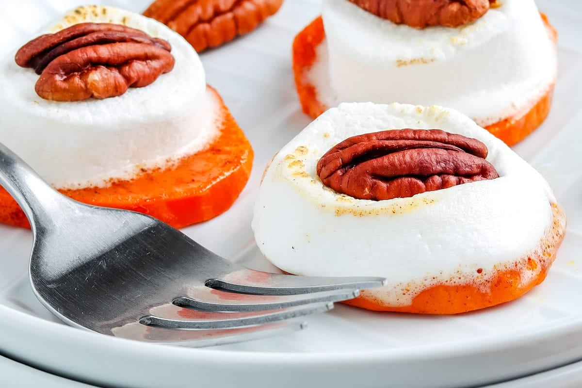 Slices of sweet potato topped with toasted marshmallow and a pecan on a white plate with a fork.