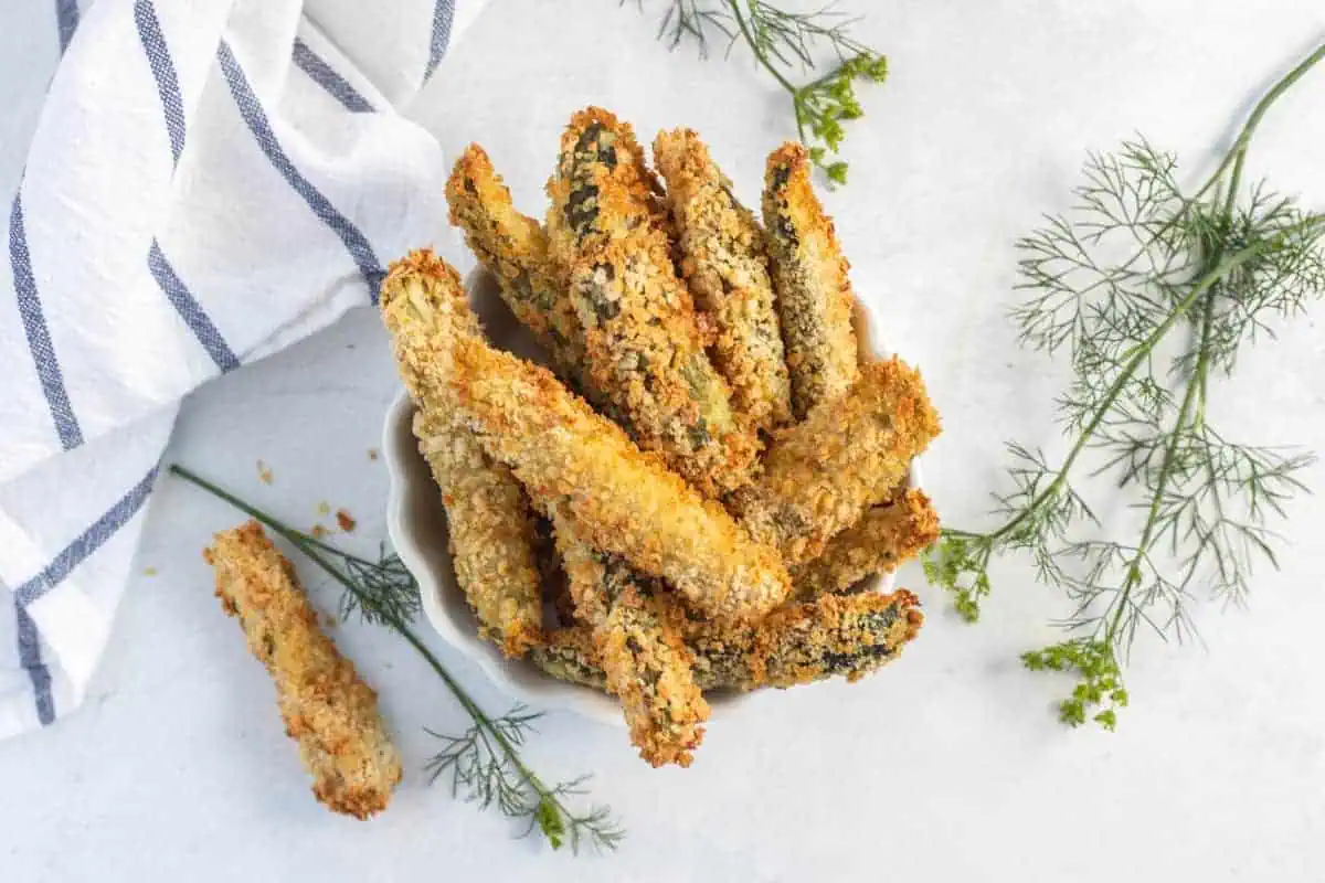A bowl of crispy breaded zucchini fries with fresh dill sprigs on a white surface.