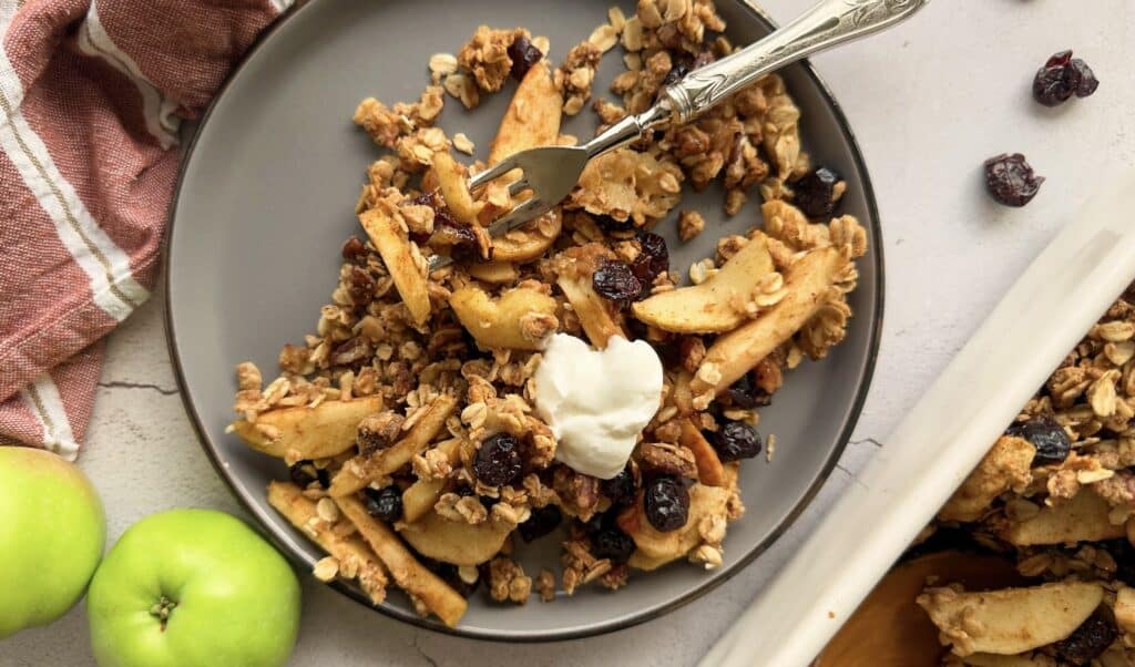 Gray plate with apple crisp, granola, dried berries, and a dollop of cream, next to two green apples.