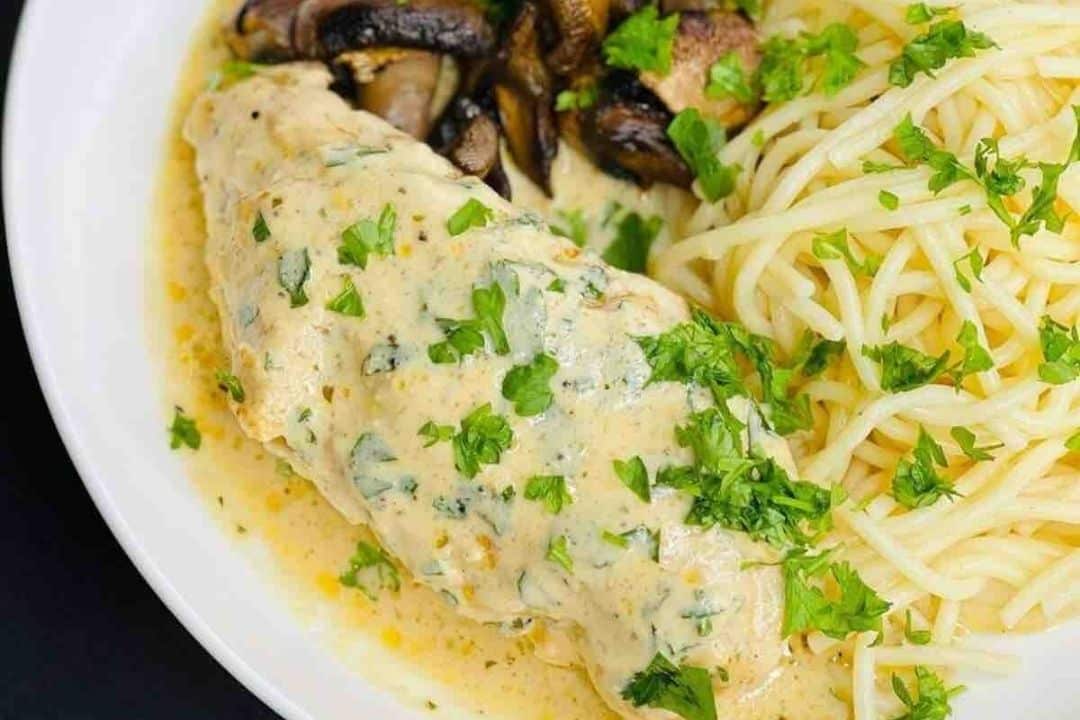 Chicken breast with creamy herb sauce, mushrooms, and spaghetti, garnished with chopped parsley on a white plate.