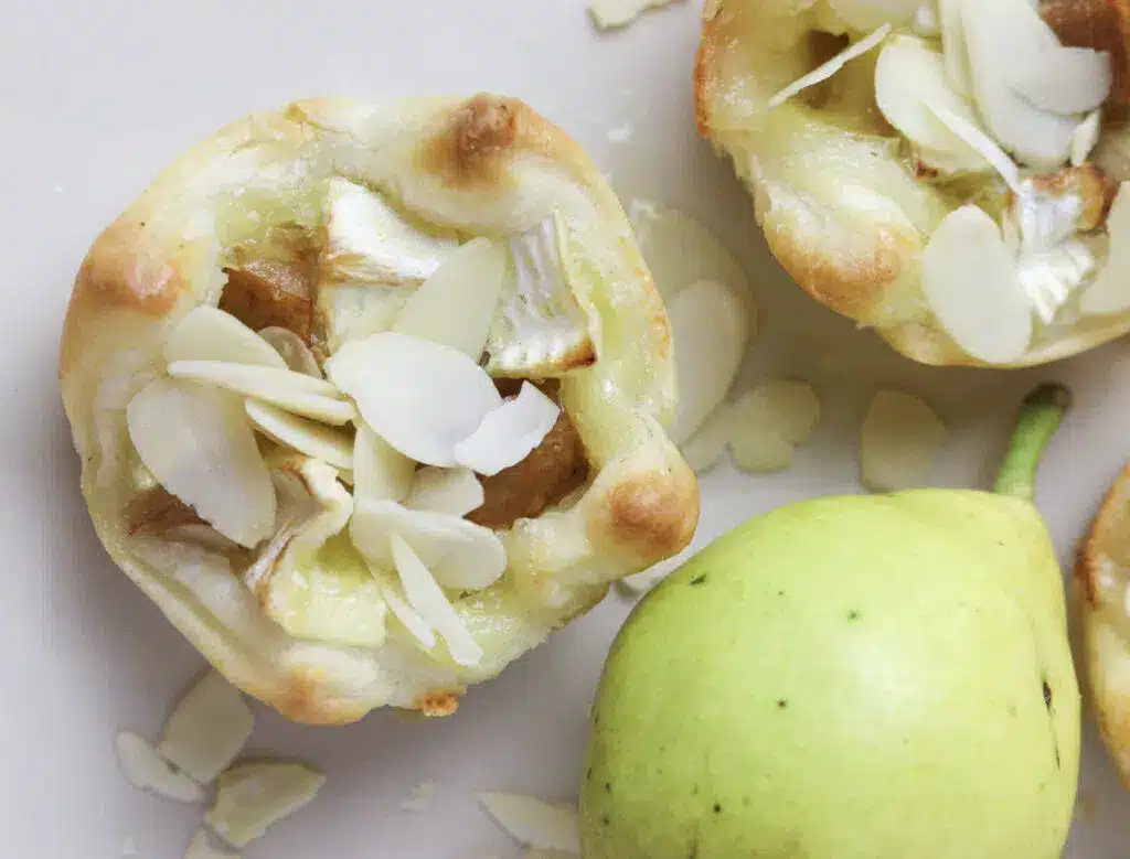 Mini tart with melted cheese, almond slices, and pear pieces next to a whole pear, on a white surface.