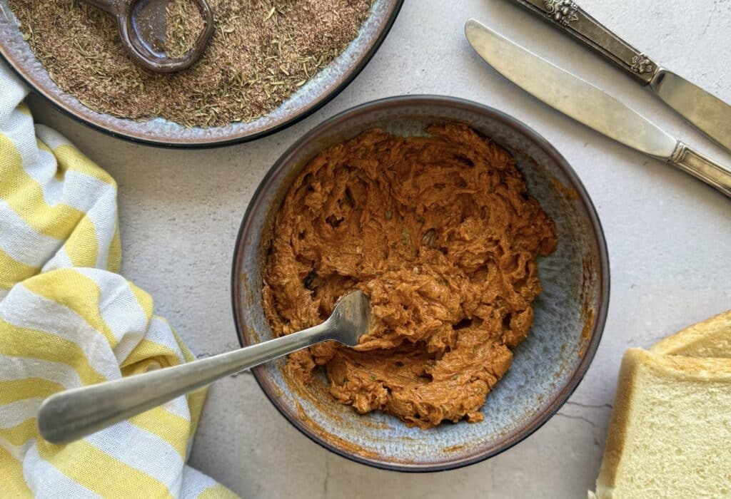 A bowl of paprika butter with a fork, next to bread, a knife, and a striped yellow towel on a table.