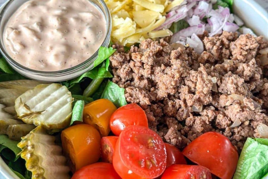Ground beef, sliced tomatoes, pickles, cheese, onions, and sauce arranged in a bowl on greens.