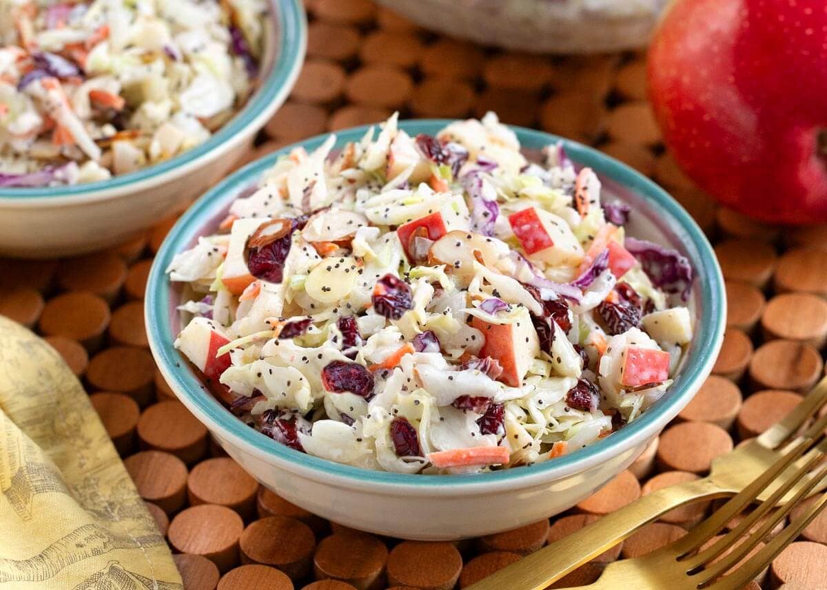 Bowl of coleslaw with chopped apples, cranberries, and poppy seeds on a wooden mat.