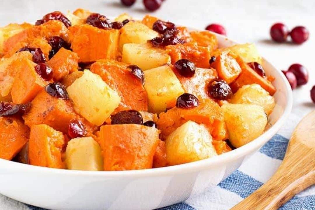 Bowl of baked sweet potatoes and pineapples topped with cranberries, with a wooden spoon beside it.