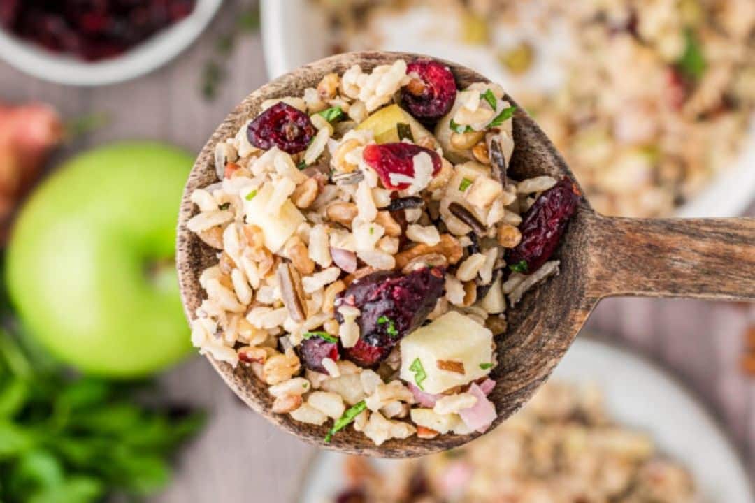 Wooden spoon holding wild rice salad with cranberries, apples, herbs, and grains.
