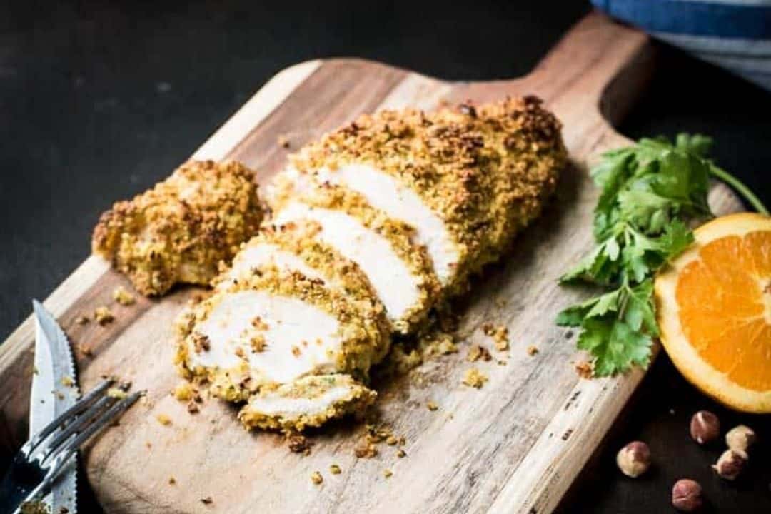 Sliced crusted chicken breast on a wooden board with coriander, orange, and nuts beside it.
