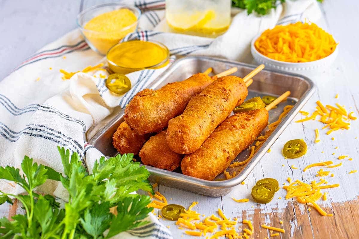 Four corn dogs on a tray with grated cheese, jalapeños, mustard, and parsley on a light table.