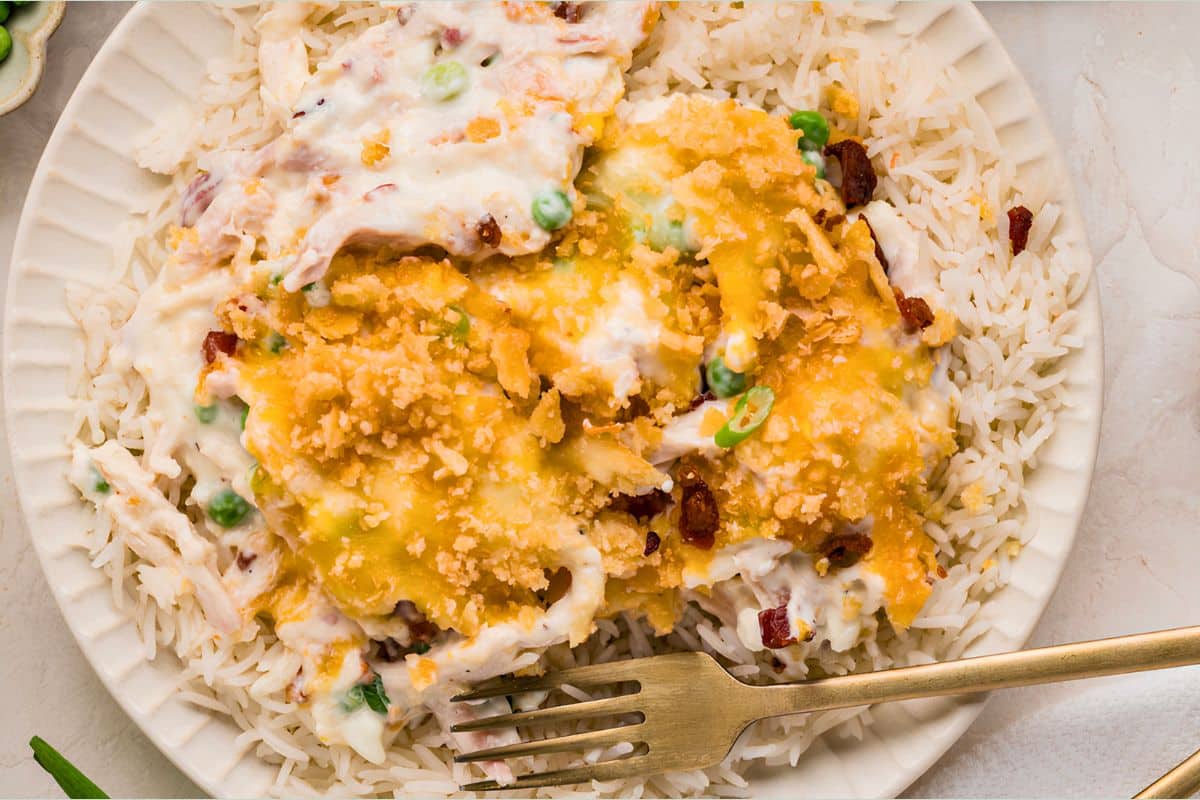 A plate of rice topped with creamy chicken casserole, melted cheese, peas, bacon, and a golden crumb topping.
