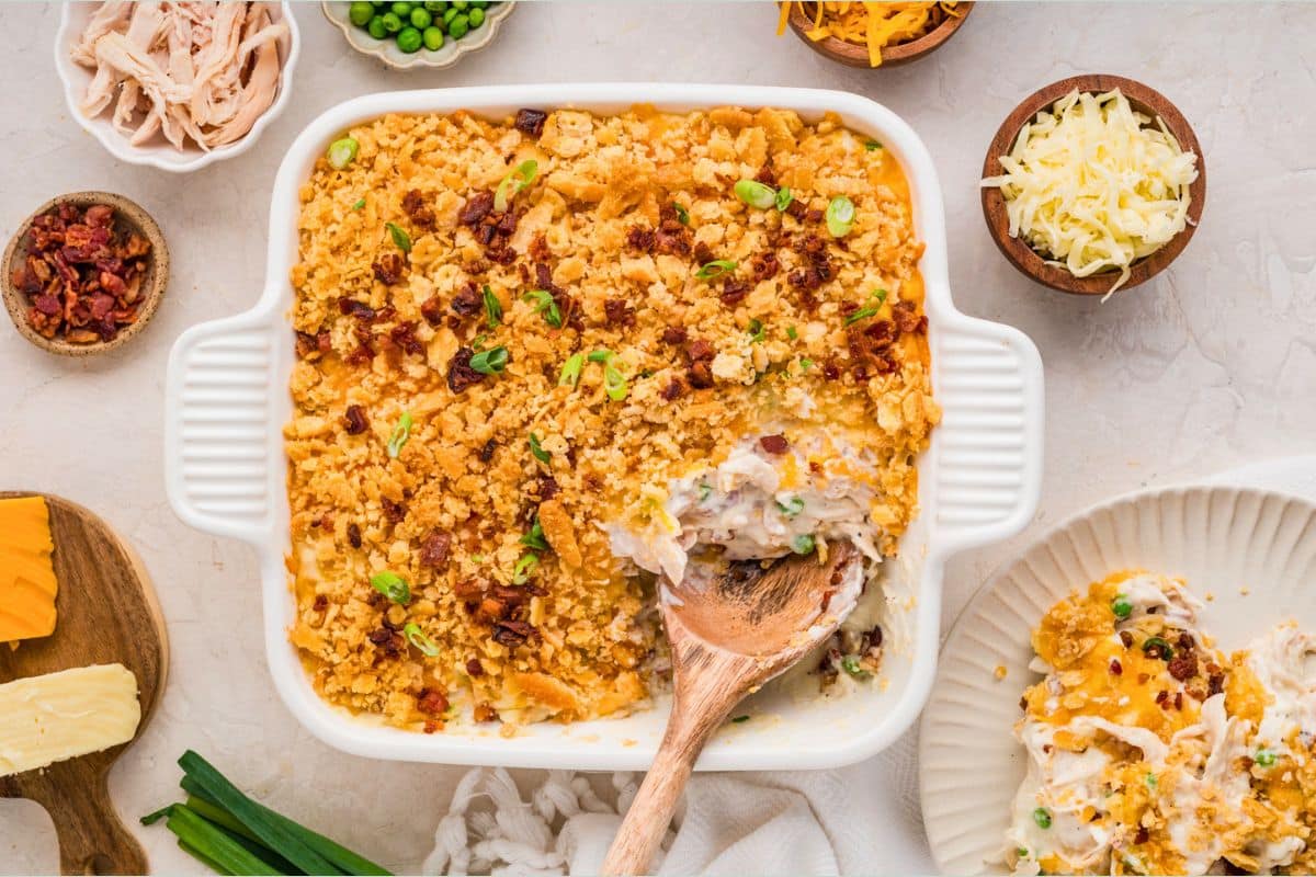 A baked casserole with a golden crumb topping, served with a wooden spoon, surrounded by cheese and toppings.