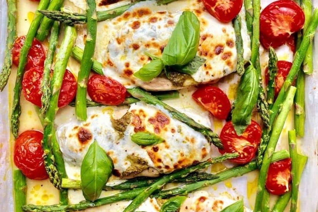 Baked chicken with melted cheese, asparagus, cherry tomatoes, and fresh basil leaves on a sheet pan.