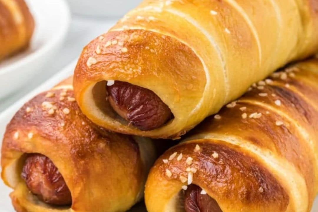 Three golden brown pigs in a blanket with sesame seeds, stacked on a white plate.