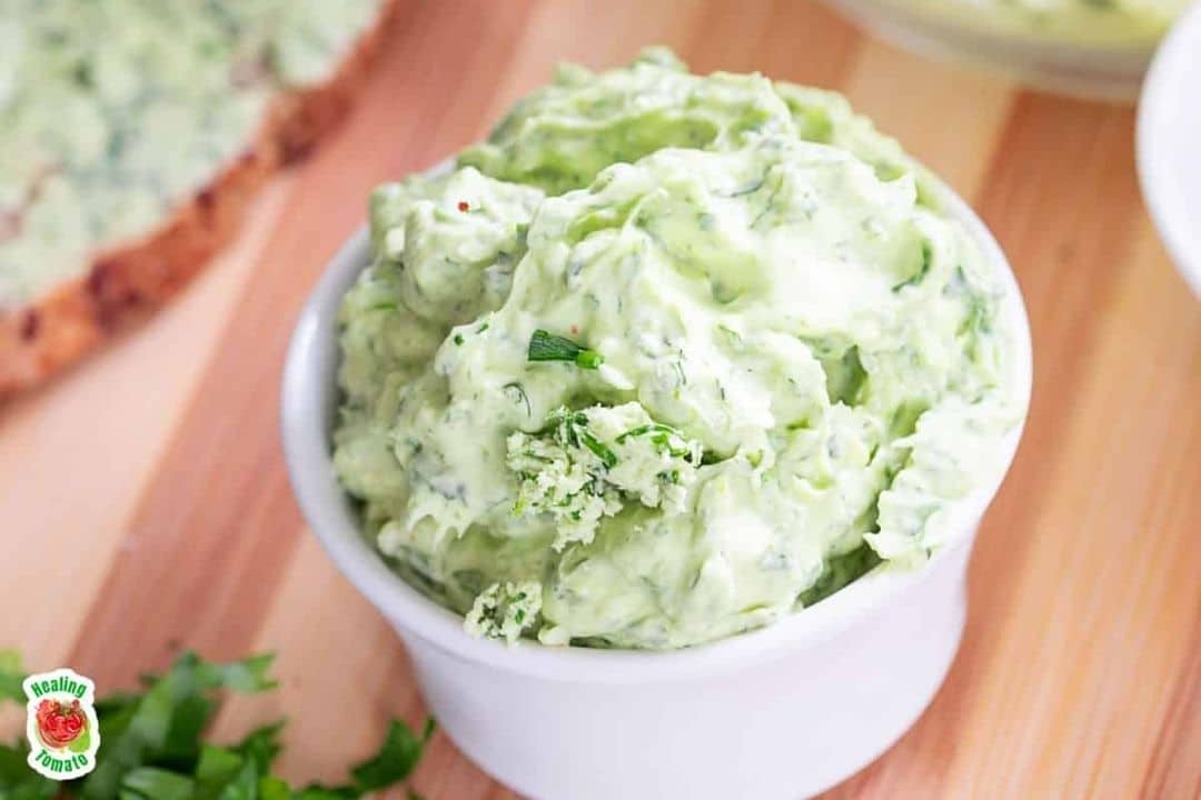 Green herb butter in a small white bowl on a wooden surface, with herbs scattered nearby.