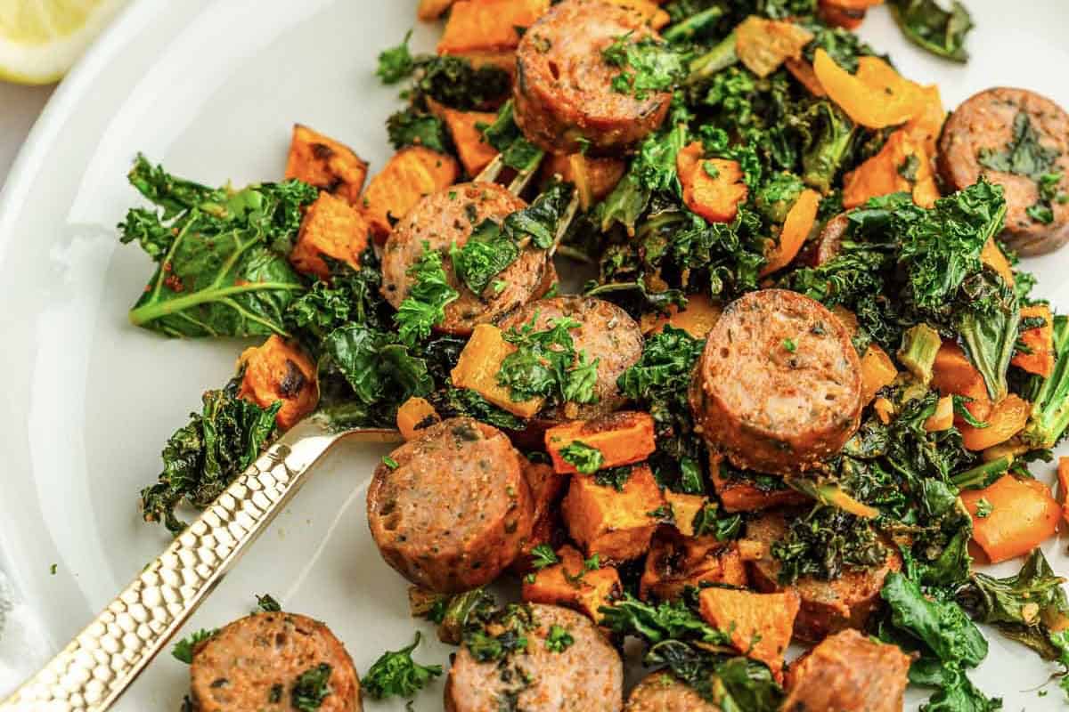 Sliced sausage, kale, and roasted sweet potatoes on a white plate with a gold fork.