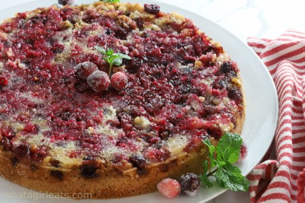 A cranberry cake topped with sugared cranberries and mint sits on a white plate next to a red-striped towel.