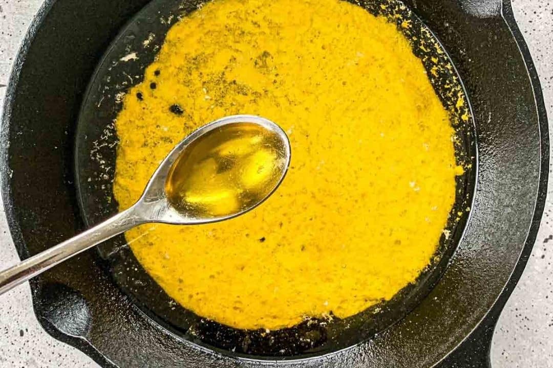 A spoon with oil held over a skillet containing a yellow spice mixture on a stovetop.