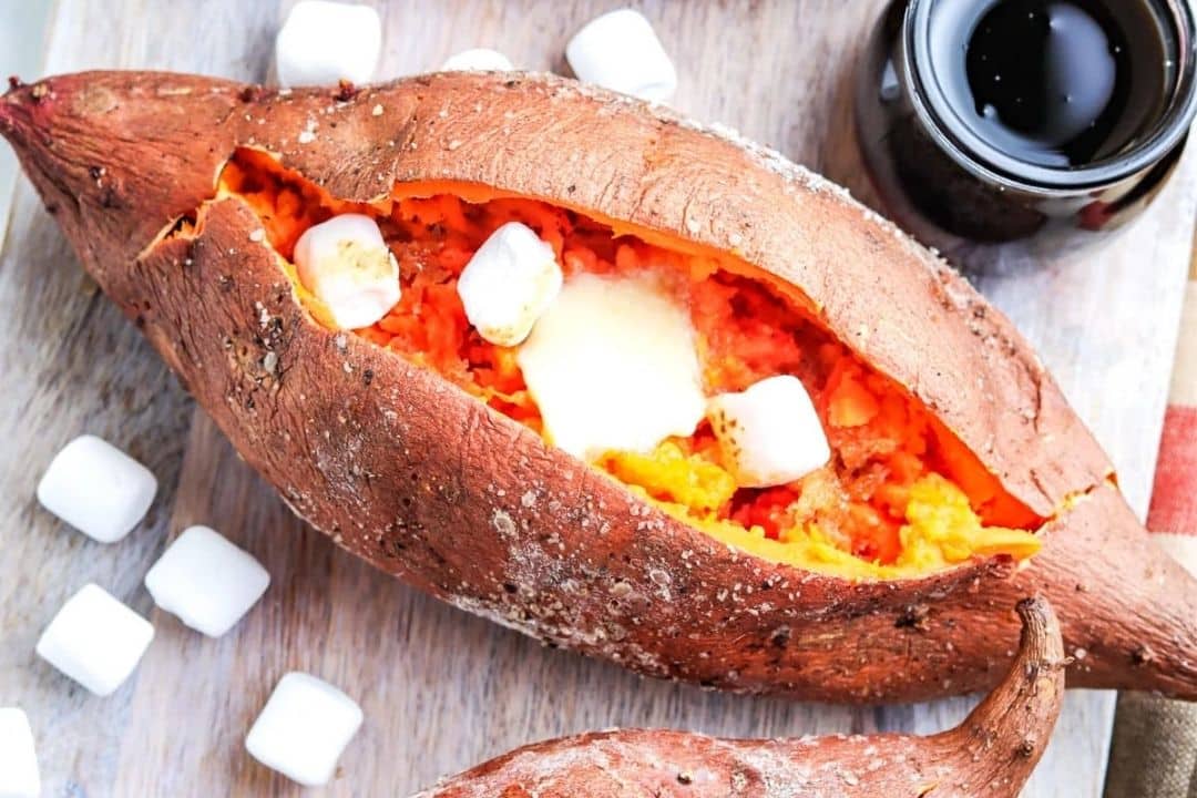 Baked sweet potato split open, filled with butter and marshmallows, on a wooden board next to syrup.