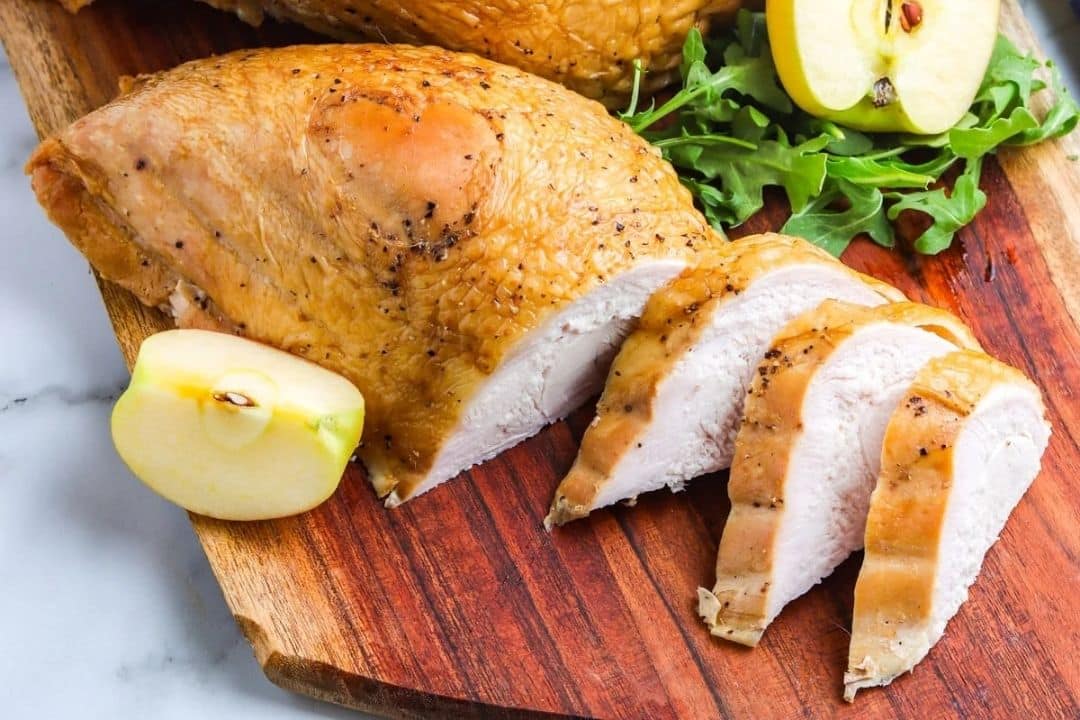 Sliced roasted turkey breast on a wooden board with apple halves and fresh arugula.