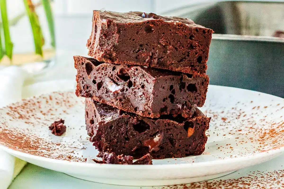 A stack of three chocolate brownies on a white plate dusted with cocoa powder.