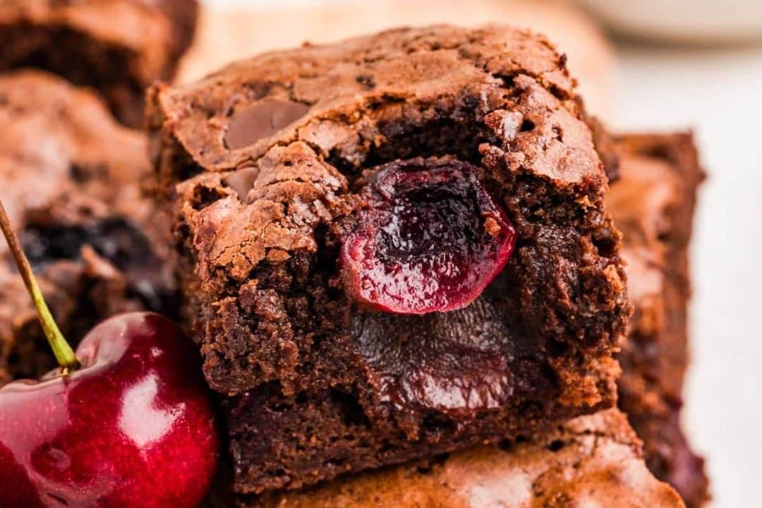 A chocolate brownie with a cherry baked into the center, next to a fresh cherry.