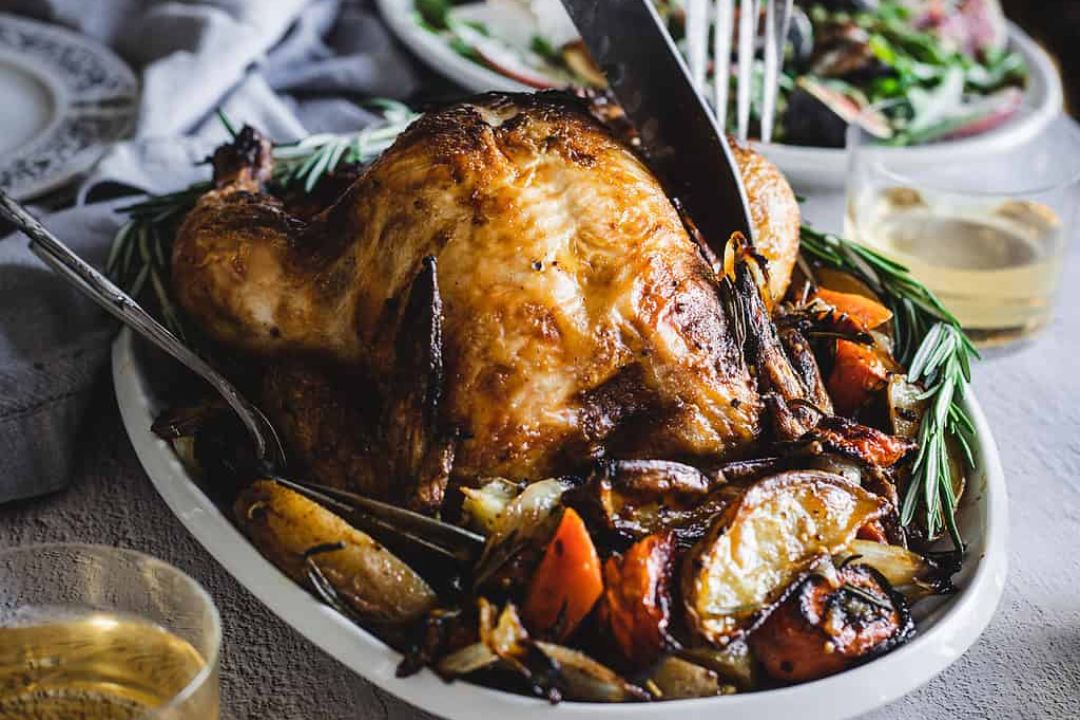 Roasted chicken on a platter with potatoes, carrots, and rosemary, being carved with a knife.