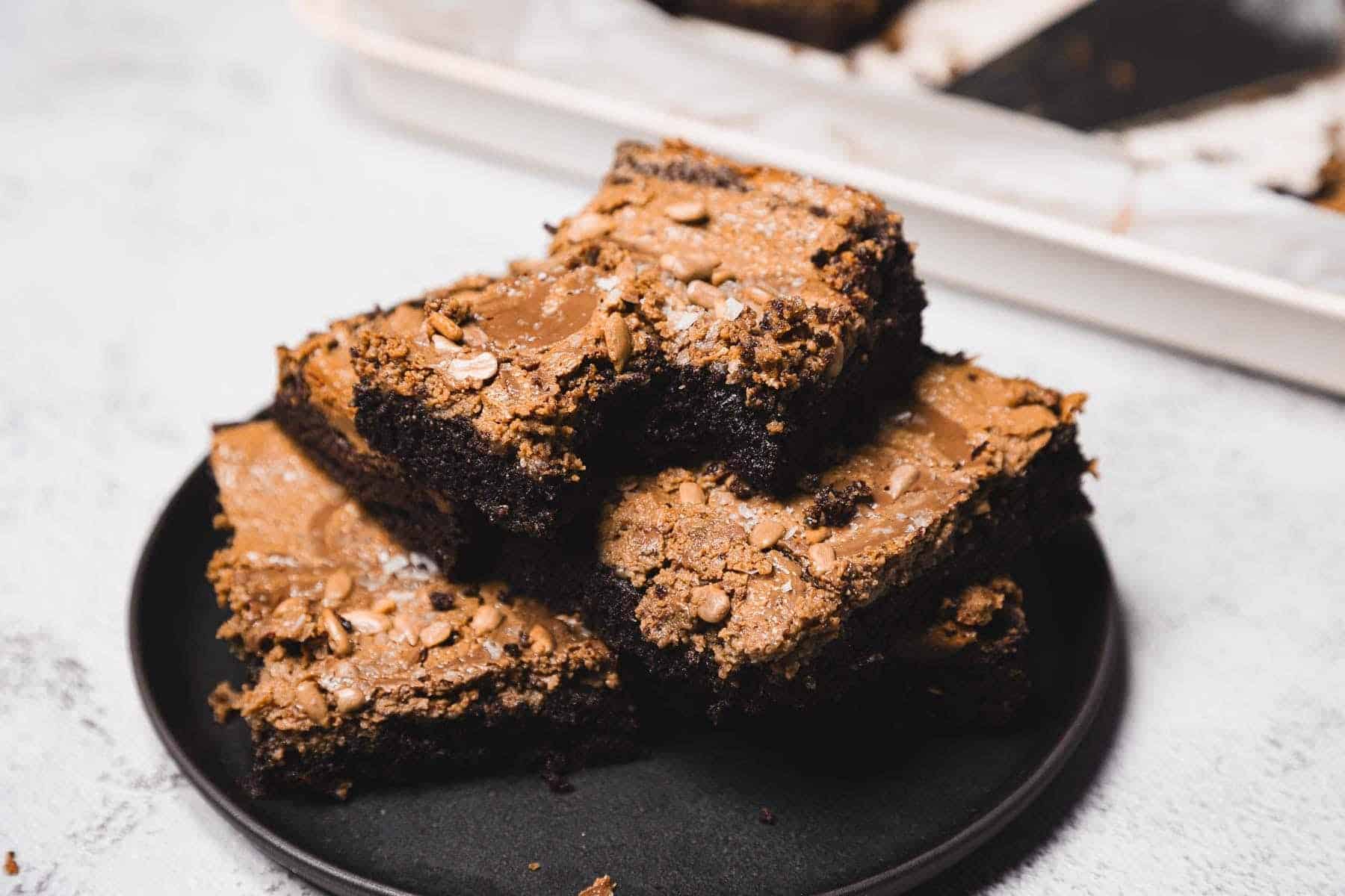 A stack of chocolate brownies topped with seeds and sea salt on a black plate.