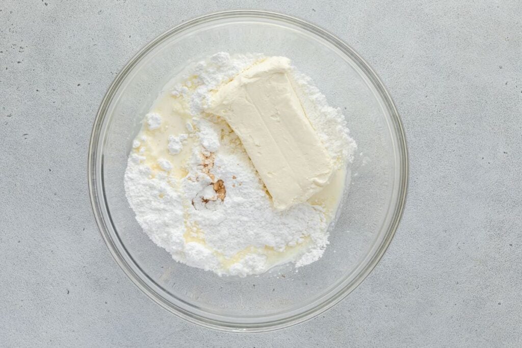 A glass bowl containing a block of cream cheese, powdered sugar, and liquid ingredients.