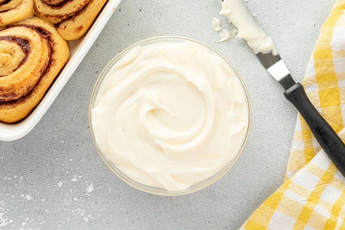 A bowl of cream cheese frosting next to cinnamon rolls and an offset spatula.
