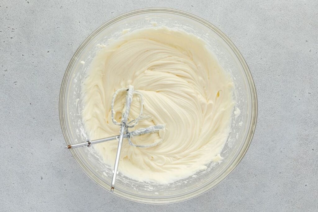 Creamy white frosting being whipped in a glass bowl with electric beaters.