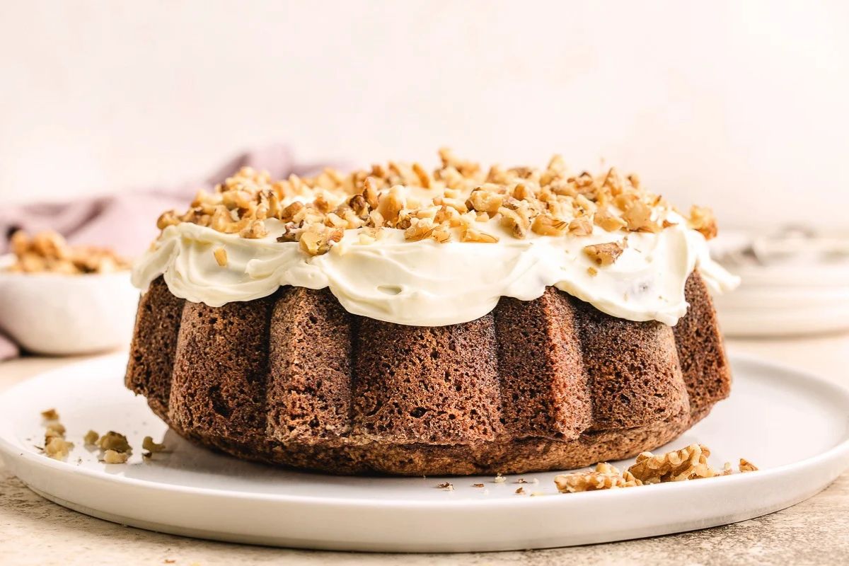 Bundt cake with white frosting topped with chopped walnuts on a white plate.