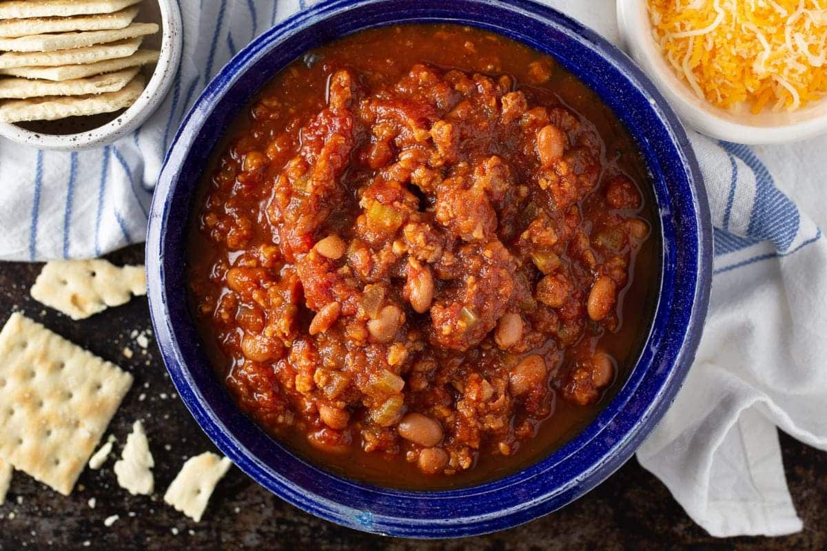 A blue bowl of chili surrounded by crackers and shredded cheese.