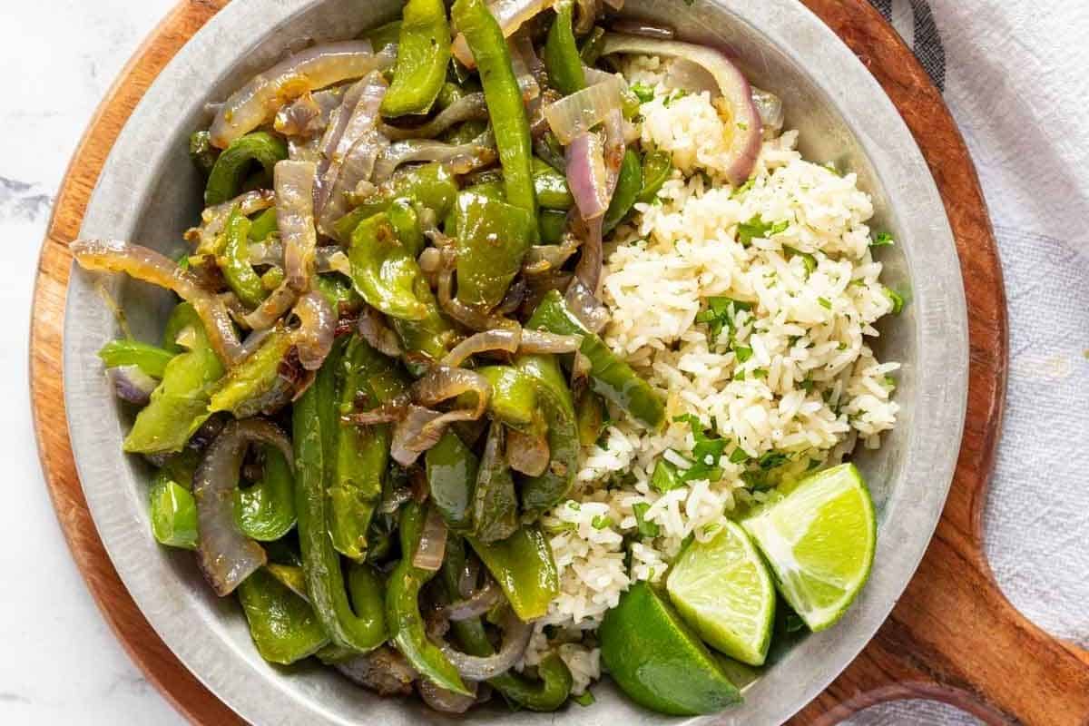 A metal bowl filled with sautéed green peppers, onions, and rice, served with two lime wedges.