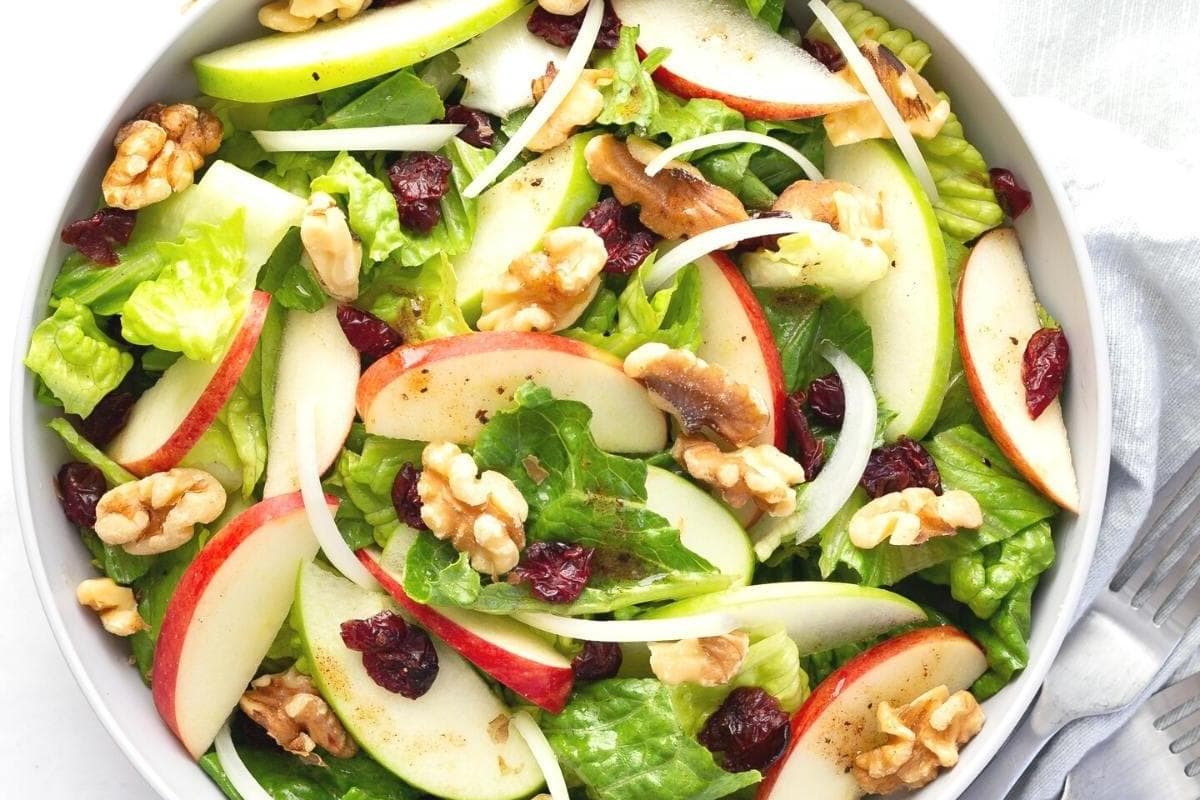 A salad with sliced apples, walnuts, and dried cranberries in a white bowl.