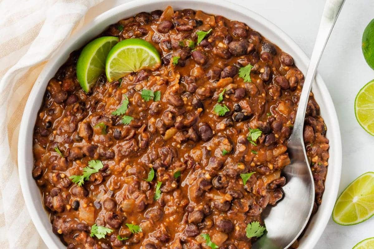 A bowl of black bean stew garnished with cilantro and lime wedges, with a serving spoon.