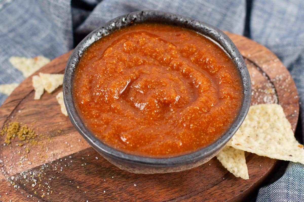 A bowl of red salsa on a wooden board with tortilla chips.